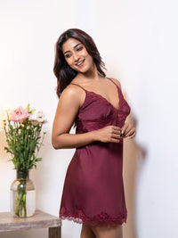 Woman wearing a burgundy nightgown with lace details, standing next to a vase of flowers on a white background.