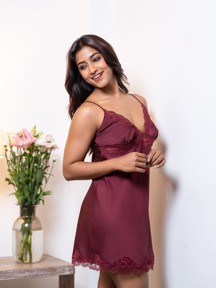 Woman wearing a burgundy nightgown with lace details, standing next to a vase of flowers on a white background.