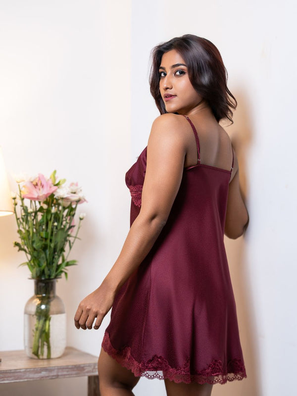 Woman wearing a burgundy lace nightgown in a softly lit room with flowers in the background.