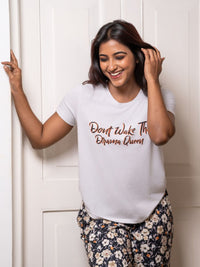 Woman wearing a white t-shirt with text, standing against a white wall.