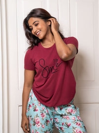 Woman wearing a maroon t-shirt with 'Smile' and floral pants against a white wall.
