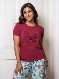 Woman wearing a maroon t-shirt with 'Smile' text and floral pants against a white background