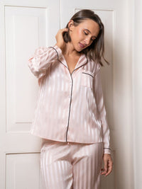Woman wearing a light pink satin pajama set with black piping against a white background