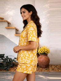 Woman wearing a yellow floral pajama set standing in a home setting with plants.