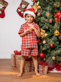 Child in red plaid pajamas and Santa hat standing in front of a decorated Christmas tree with presents.