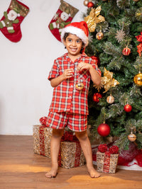 Child in red plaid pajamas and Santa hat standing in front of a decorated Christmas tree with stockings.