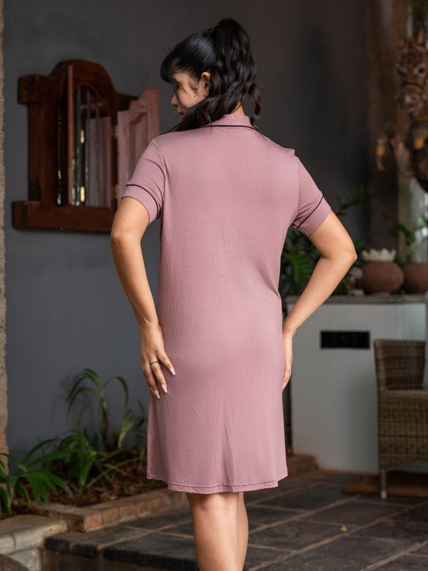 Woman wearing a mauve dress standing in an outdoor setting with plants and a wall in the background.