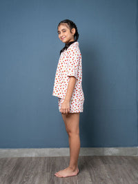 Young girl wearing a white shirt with red patterns and matching shorts against a blue wall.