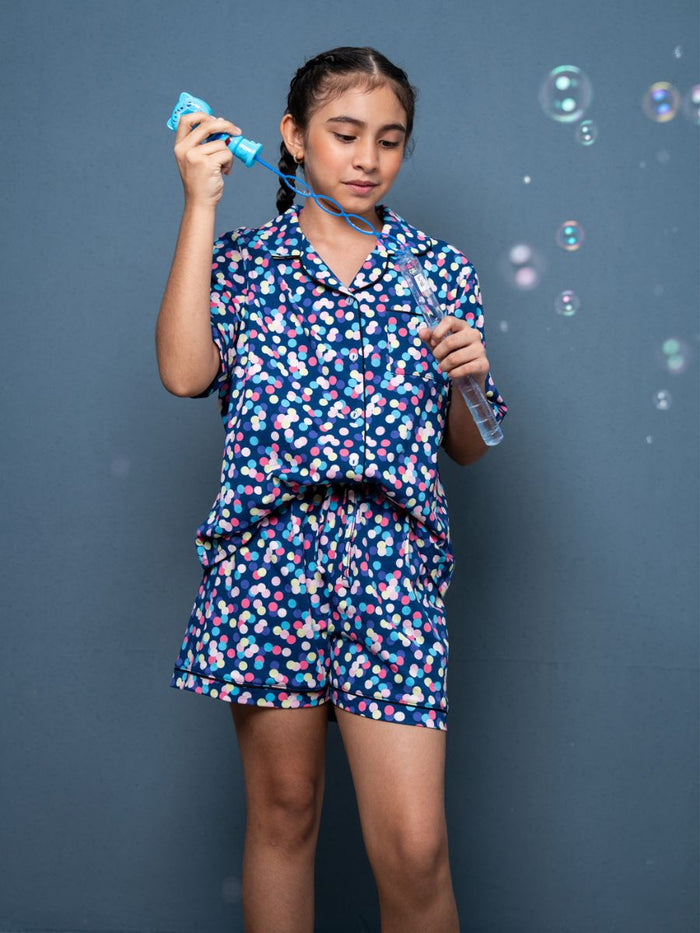 Girl in a colorful polka dot outfit blowing bubbles against a gray background