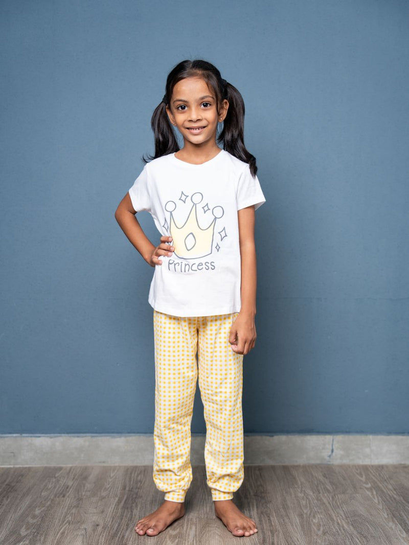 Young girl wearing a 'Princess' t-shirt and yellow checkered pants against a blue wall.