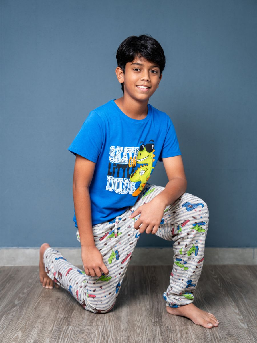 Child wearing a blue t-shirt with a graphic and patterned pants, sitting on the floor against a gray wall.