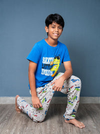 Child wearing a blue t-shirt with a graphic and patterned pants, sitting on the floor against a gray wall.