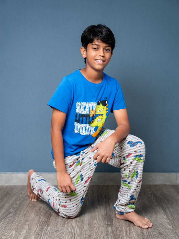 Child wearing a blue t-shirt with a graphic and patterned pants, sitting on the floor against a gray wall.