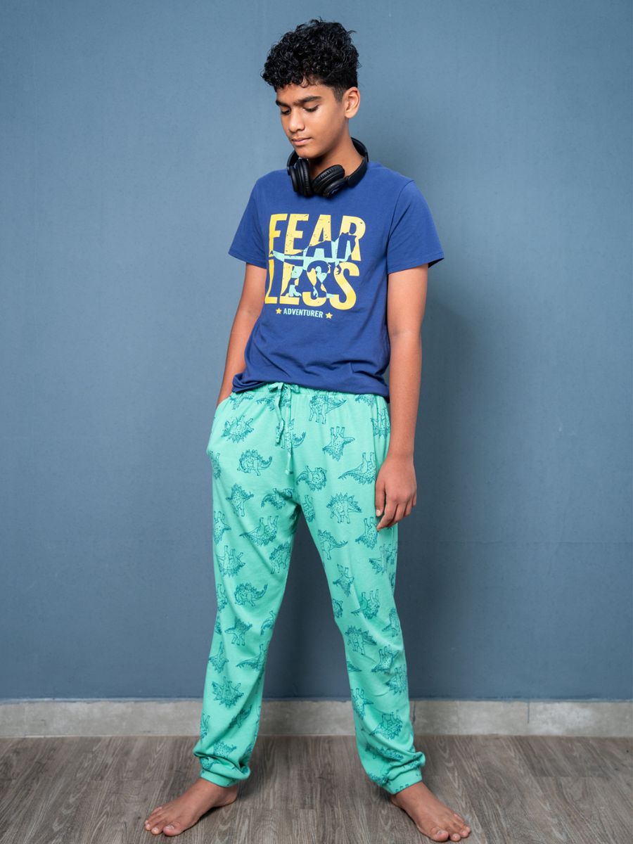 Person wearing a blue t-shirt with 'FEAR LESS' text and green patterned pants against a gray wall.