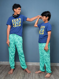 Two children wearing matching blue t-shirts and green pants against a gray wall.