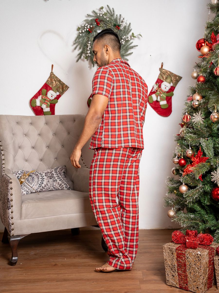 Man in red plaid pajamas standing in a room decorated for Christmas with a tree and stockings.