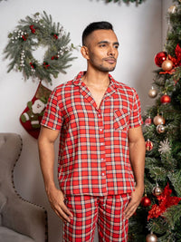 Man wearing red plaid pajamas standing in front of a decorated Christmas tree and wreath.