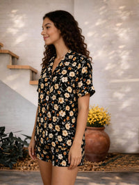 Woman wearing a black floral dress standing in a home setting with plants and a staircase.