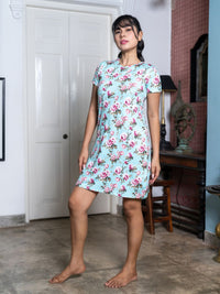 Woman wearing a floral dress standing in a room with a white door and wooden furniture.