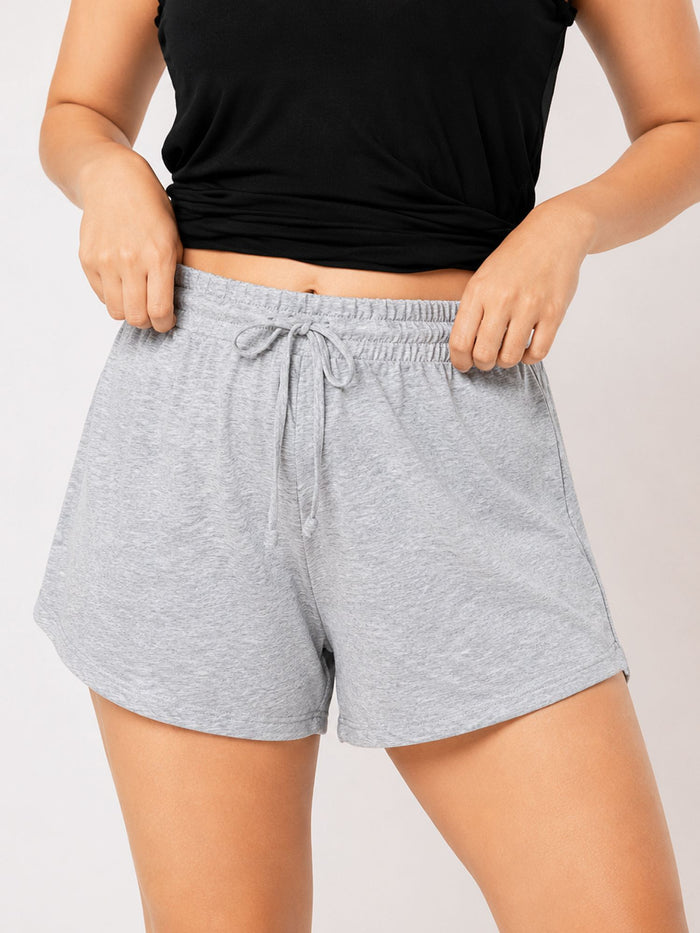 A person wearing light heather gray drawstring shorts and a black cropped tank top, seated against a plain light gray studio background.