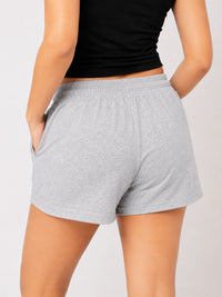 A person wearing light heather gray drawstring shorts and a black cropped tank top, seated against a plain light gray studio background.
