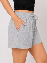 A close-up of a person wearing light heather gray drawstring shorts with side pockets, paired with a black cropped top, against a plain light gray studio background.