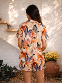 Woman wearing a floral outfit standing outdoors with plants and a white wall in the background