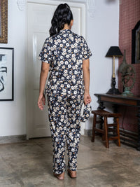 Woman wearing a floral outfit standing in a room with a lamp and framed pictures on the wall.