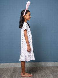 Young girl wearing a white dress with a pattern and bunny ears against a blue wall.