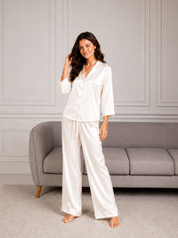 Woman wearing white satin pajamas standing in a room with a gray sofa.