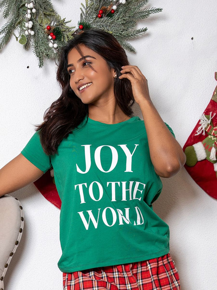 Woman wearing a green 'JOY TO THE WORLD' shirt with Christmas decorations in the background