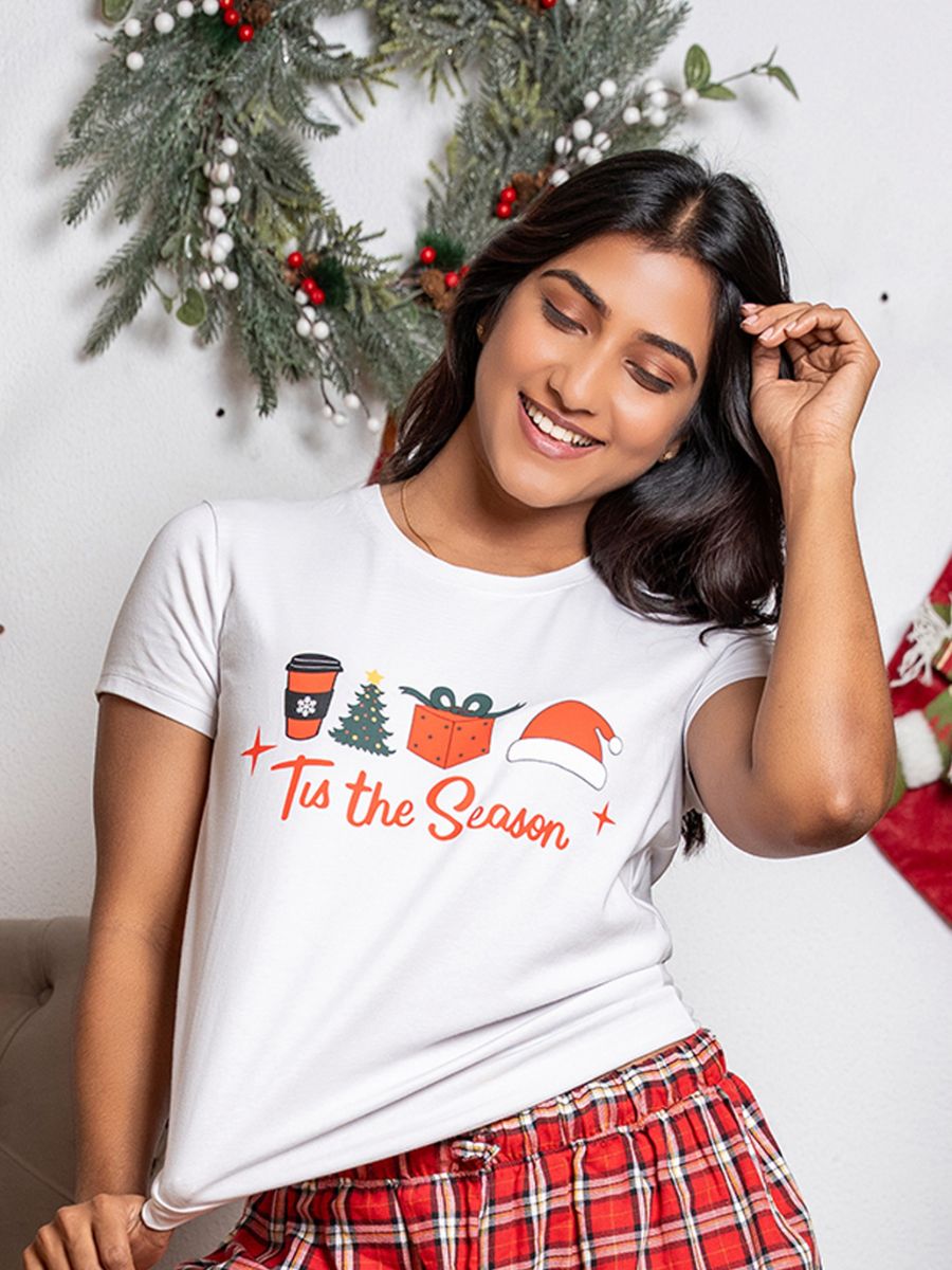 Woman wearing a white t-shirt with Christmas-themed graphics and text, standing in front of a decorated wall.