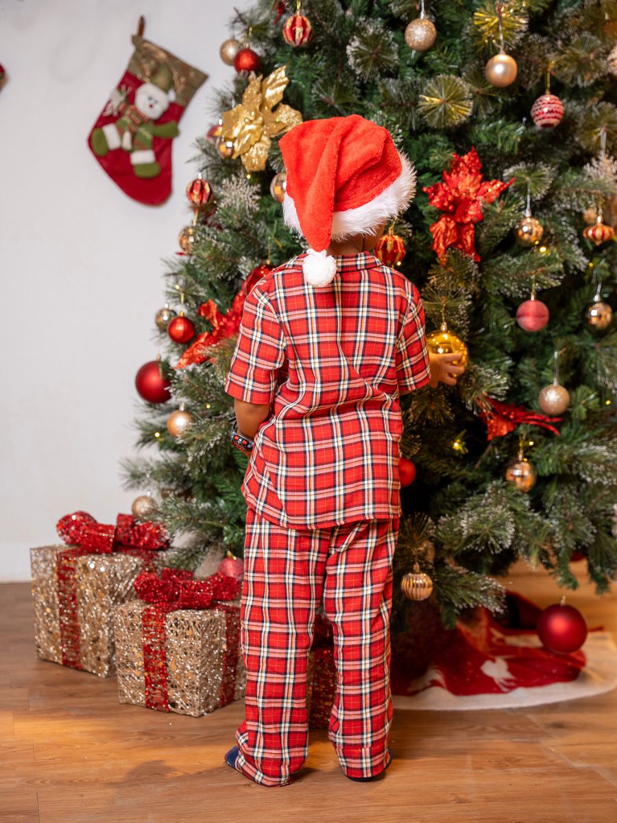 Child in red plaid pajamas and Santa hat standing in front of a decorated Christmas tree.