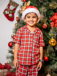 Child wearing a red plaid outfit and Santa hat in front of a decorated Christmas tree.