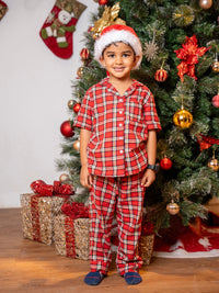 Child in red plaid pajamas and Santa hat standing in front of a decorated Christmas tree with presents.
