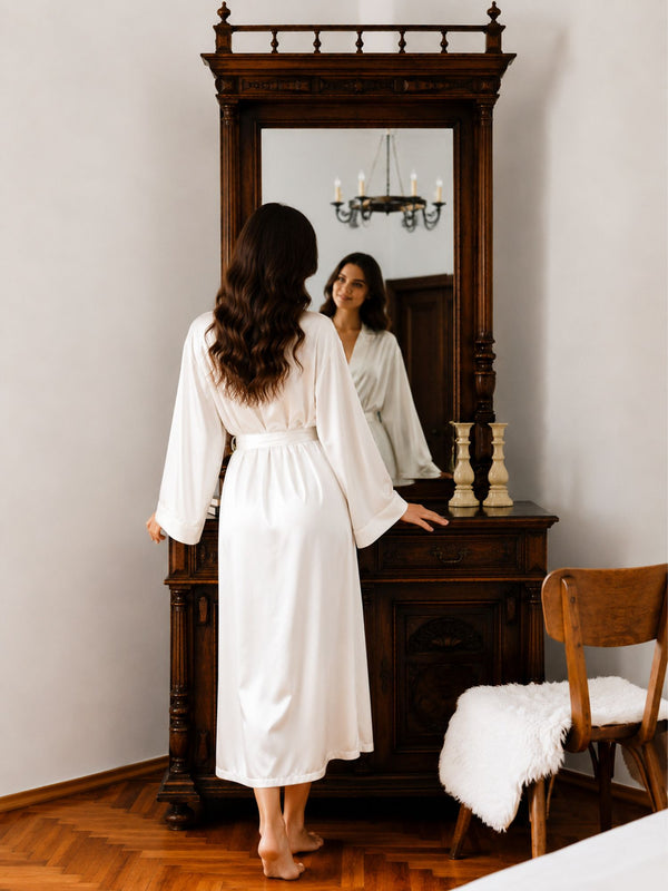 Woman in a white robe standing in front of a large mirror in a room with wooden furniture.