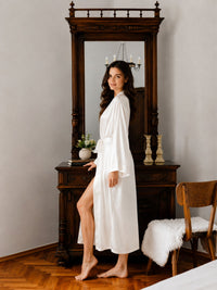Woman in a white robe standing in a room with wooden furniture and a mirror.