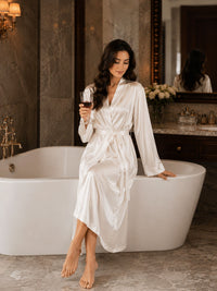 Woman in a white satin robe holding a glass of red wine in a bathroom.