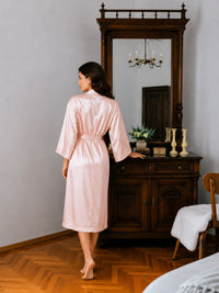 Woman in a pink robe standing in a room with wooden furniture and a mirror.