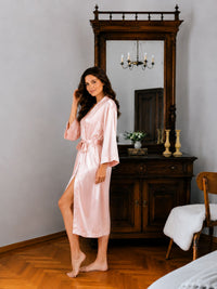 Woman in a pink satin robe standing in a bedroom with wooden furniture and a mirror.